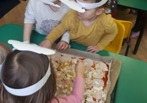 Przedszkolaki ozdabiają pizzę czerwoną papryką pokrojoną w paski.